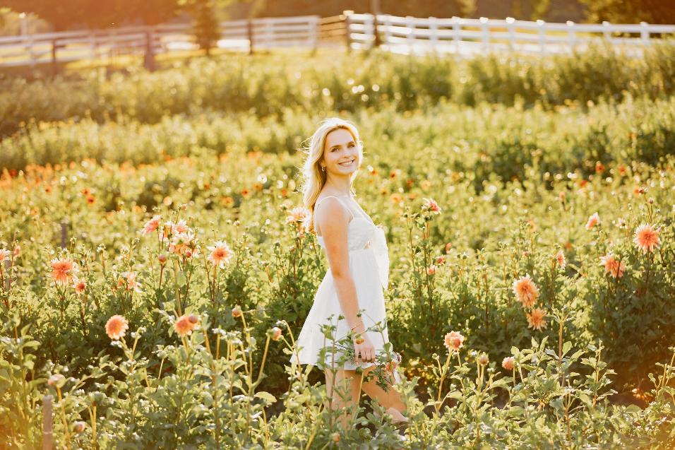 Madelyn Wright smiling over her shoulder in a sunlit field of peach dahlias, white fence in the distance