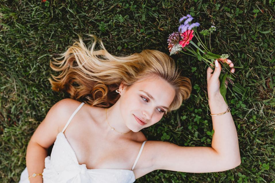 Madelyn Wright lying in the grass with her hair fanned out, smiling and holding a small bouquet of wildflowers