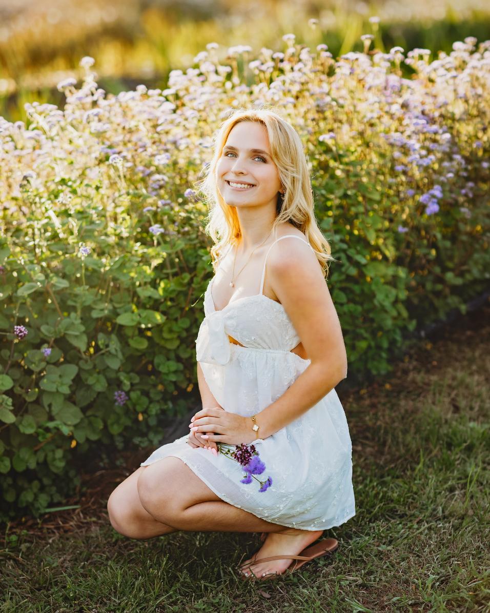 Madelyn Wright kneeling in a white dress in front of a hedge of lavender wildflowers, holding a few purple blooms